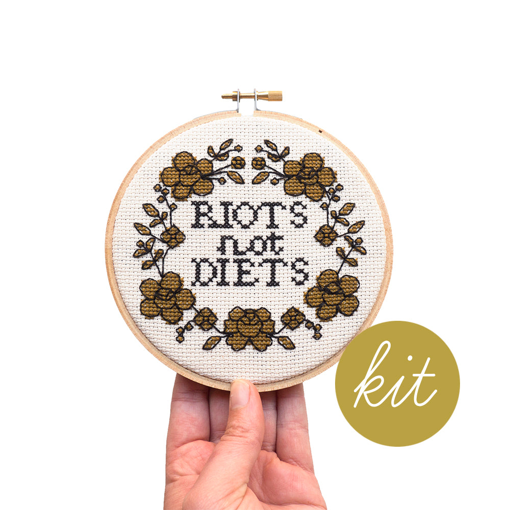 gold flowers outlined with black thread and text reading Riots not Diets in black, DIY cross stitch kit