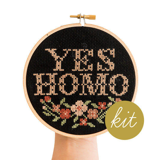 traditional cross stitch font reads Yes Homo in pink with multiple color pink flowers stitched on black aida cloth, DIY cross stitch kit
